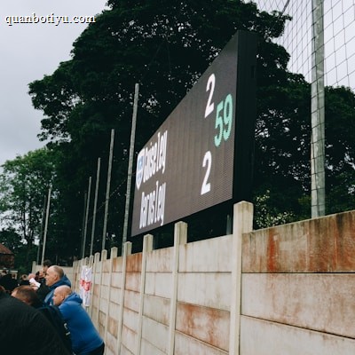 A large digital scoreboard at an outdoor sports venue displays a score of 2-2. Spectators are gathered, observing the event with interest. The scene is enclosed by a tall metal fence and surrounded by lush green trees, indicating a natural setting. Overcast skies add a muted tone to the environment.