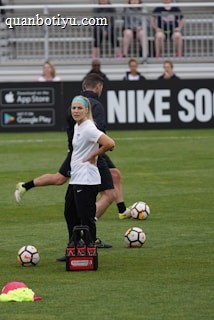 A person in sports attire stands on a soccer field, surrounded by several soccer balls and a carrier with bottles. The field is bordered by advertisements for Nike Soccer and app stores. Several people sit in bleachers in the background.
