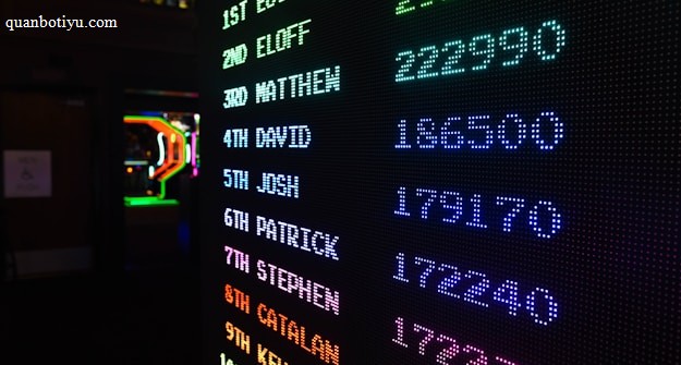 A digital leaderboard displaying names and scores in vibrant colors like green, blue, purple, and orange. The display is in a dark setting, possibly an arcade, and features a bright LED design.
