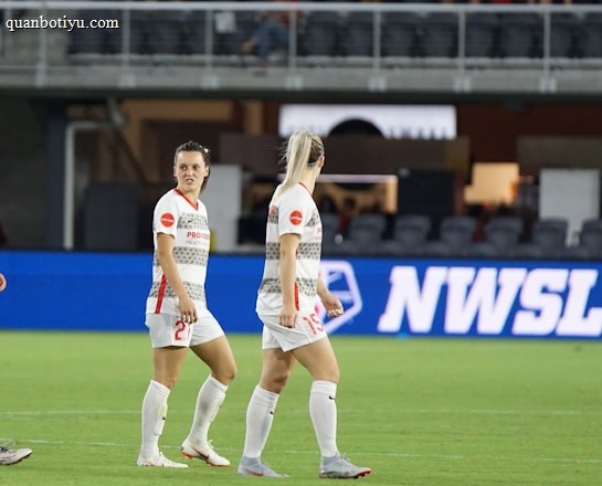 Soccer players in team uniforms stand on a grass field with a large digital advertisement screen displaying the acronym 'NWSL' in the background. The setting appears to be a stadium with rows of empty seats visible above the screen.