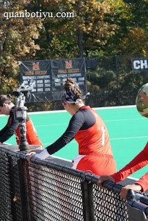 A group of athletes wearing orange uniforms are on the sideline of a sports field, resting against a fence. Behind them, a large digital scoreboard displays team names and scores. The field is bright green, and several banners with sponsor logos and championship wins are visible. The athletes appear to be taking a break, possibly before or during a field hockey game.