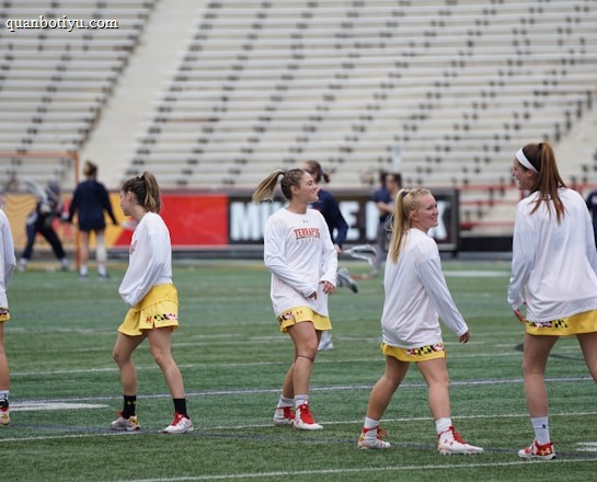 A group of young women wearing athletic gear are on a sports field. They are dressed in white jerseys with yellow shorts, featuring a distinctive design at the hem. The field has a green turf surface and the background shows empty bleachers. Some additional figures are visible in the distance, possibly referees or other players.